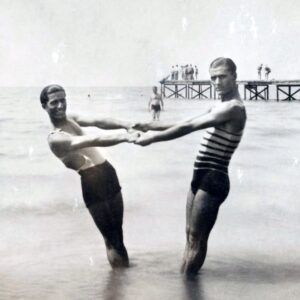 Deux hommes se tiennent par la main et regardent l'objectif en souriant. Ils portent des maillots de bain de marins. Ils ont les pieds dans la mer.